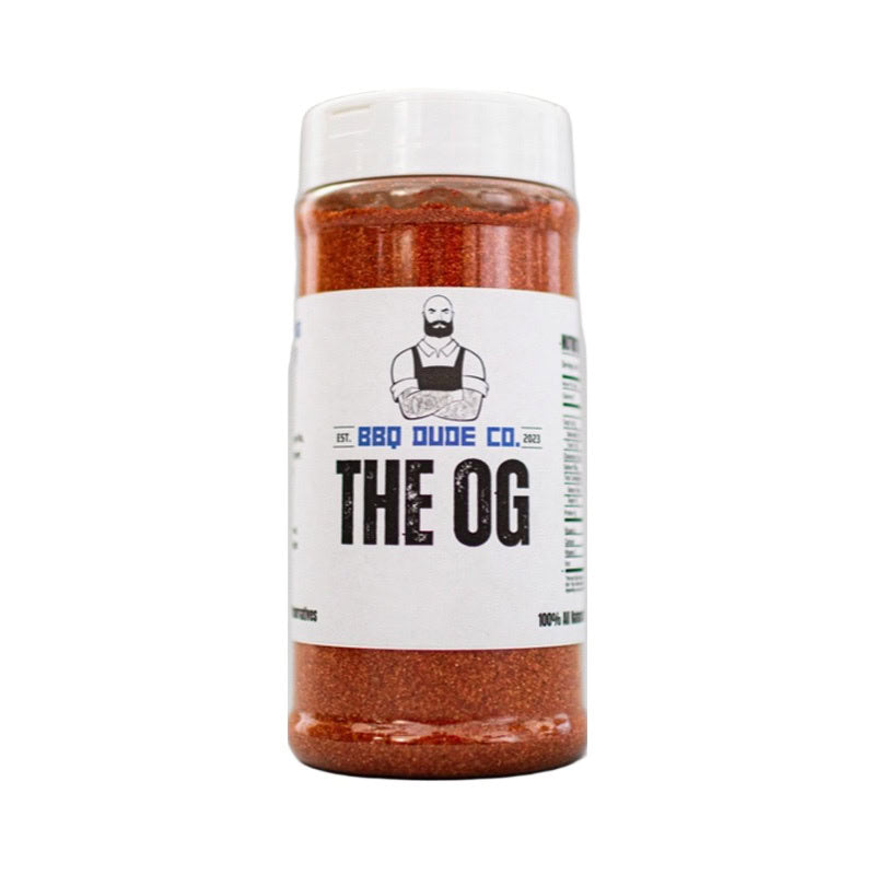 BBQ Dude The OG BBQ Seasoning bottle on wooden surface beside grilled ribs.