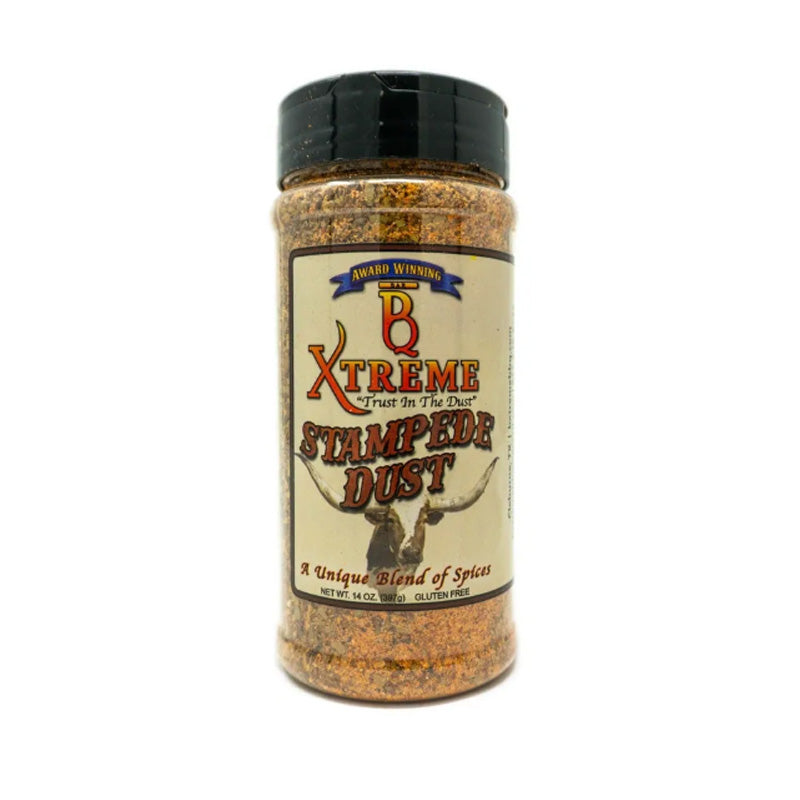 Bottle of B Xtreme BBQ Stampede Dust surrounded by grilled steaks, ribs, and peppers on a wooden board