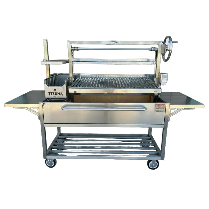 A stainless steel outdoor grill cart with a side table, lower storage shelf, and wheeled base, photographed outdoors in front of a gray brick building. The Tizona brand name is visible on the grill and side container.